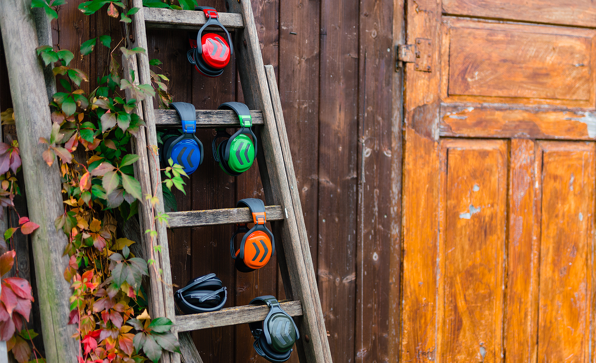 Six protections auditives color�es suspendues � une vieille �chelle en bois devant une fa�ade en bois recouverte de feuillage automnal.