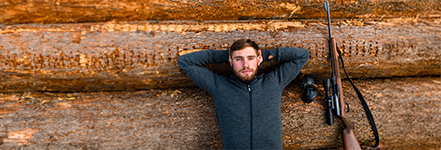 Un homme est allong� d�tendu, les bras crois�s derri�re la t�te, sur des troncs d’arbres empil�s ; � c�t� de lui se trouve un fusil de chasse avec une lunette.