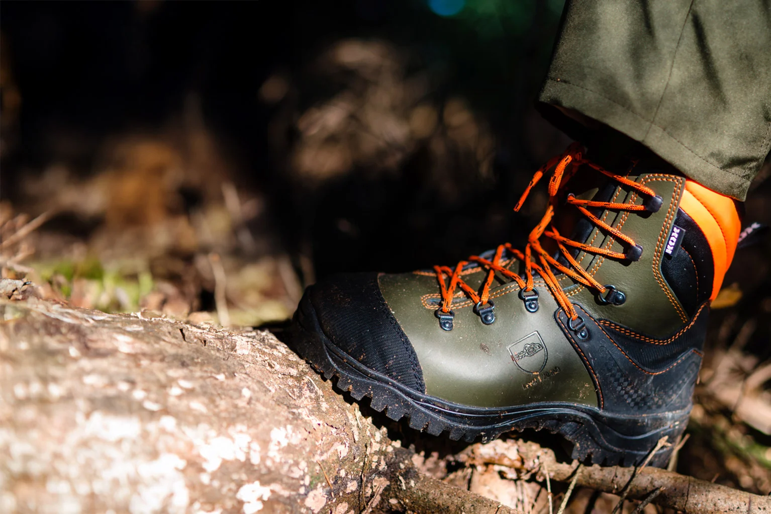 Gros plan d’une botte foresti�re robuste en vert et noir avec des lacets orange. La chaussure repose sur un tronc d’arbre en for�t, avec le sol forestier et des branches visibles de mani�re floue � l’arri�re-plan.
