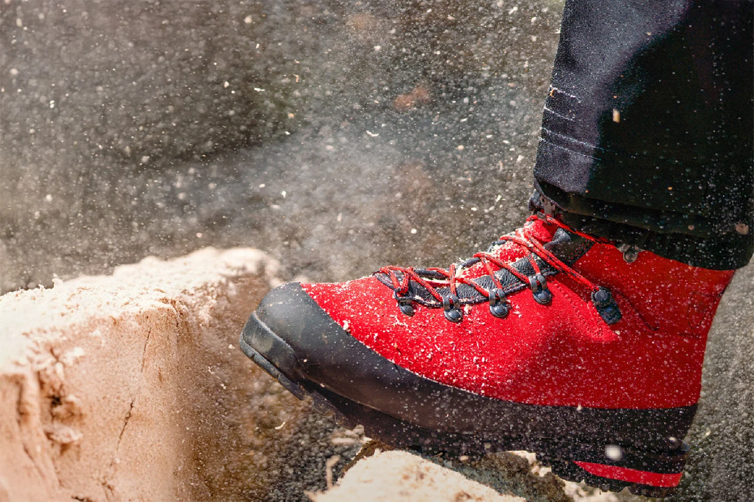 Gros plan d’une botte foresti�re rouge avec embout de protection noir et la�age. La chaussure repose sur une souche fra�chement sci�e, des copeaux de bois volent dans l’air et soulignent la situation de travail en for�t.