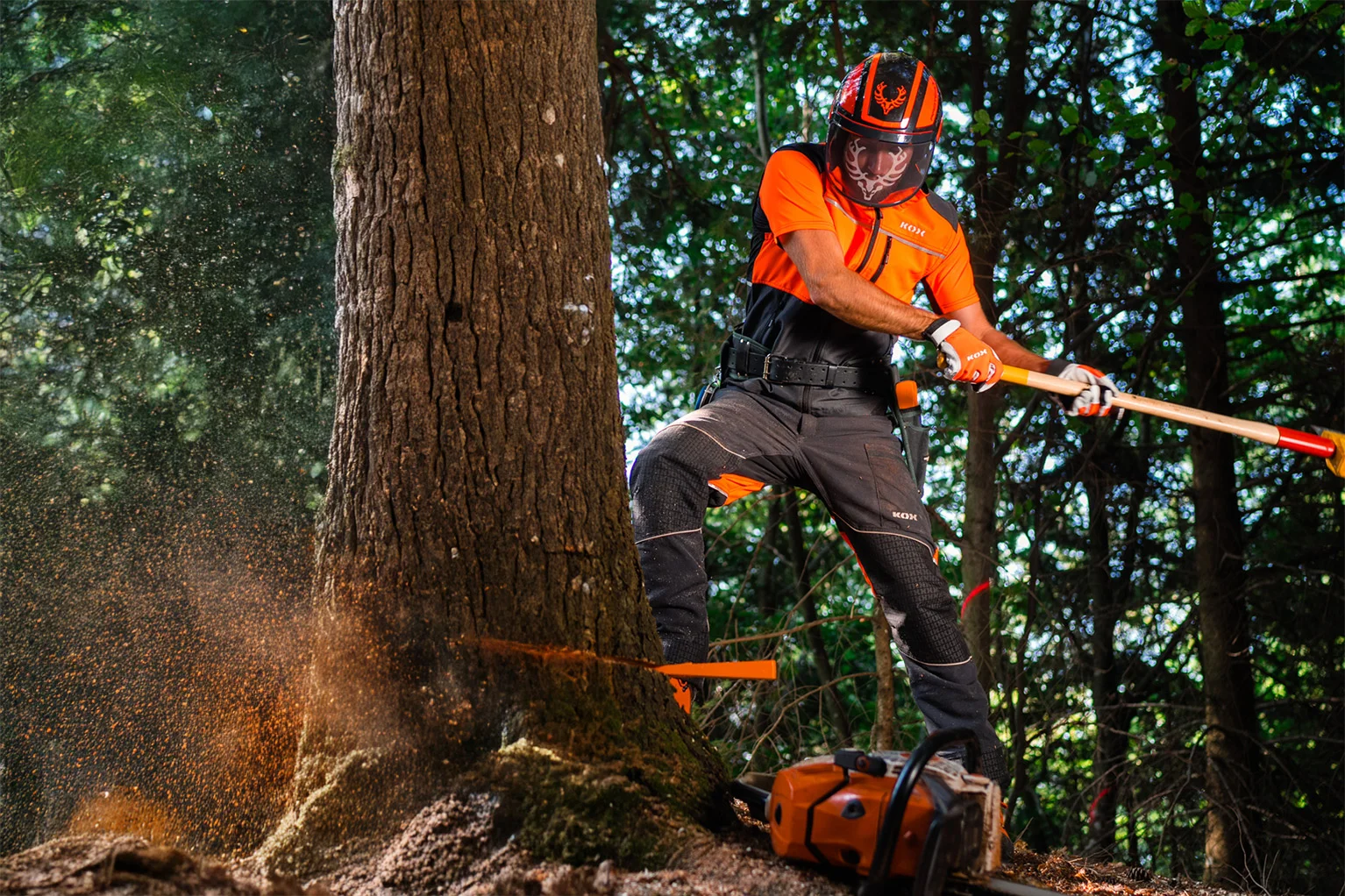 Une personne portant des v�tements de protection orange et noirs ainsi qu’un casque travaille dans la for�t sur un tronc d’arbre. Elle enfonce un coin dans le tronc � l’aide d’un levier d’abattage, des copeaux de bois volent, une tron�onneuse est pos�e au sol � c�t�.