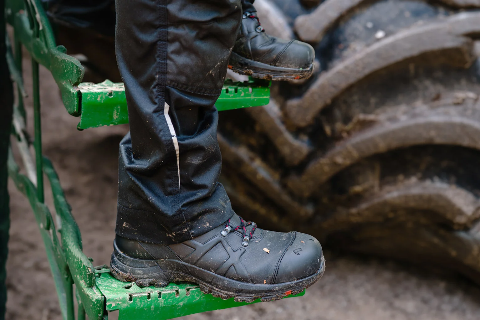 Gros plan de chaussures de travail noires avec lacets rouges pos�es sur une marche m�tallique verte ; � l’arri�re-plan, on voit un grand pneu de machine fortement encrass�.