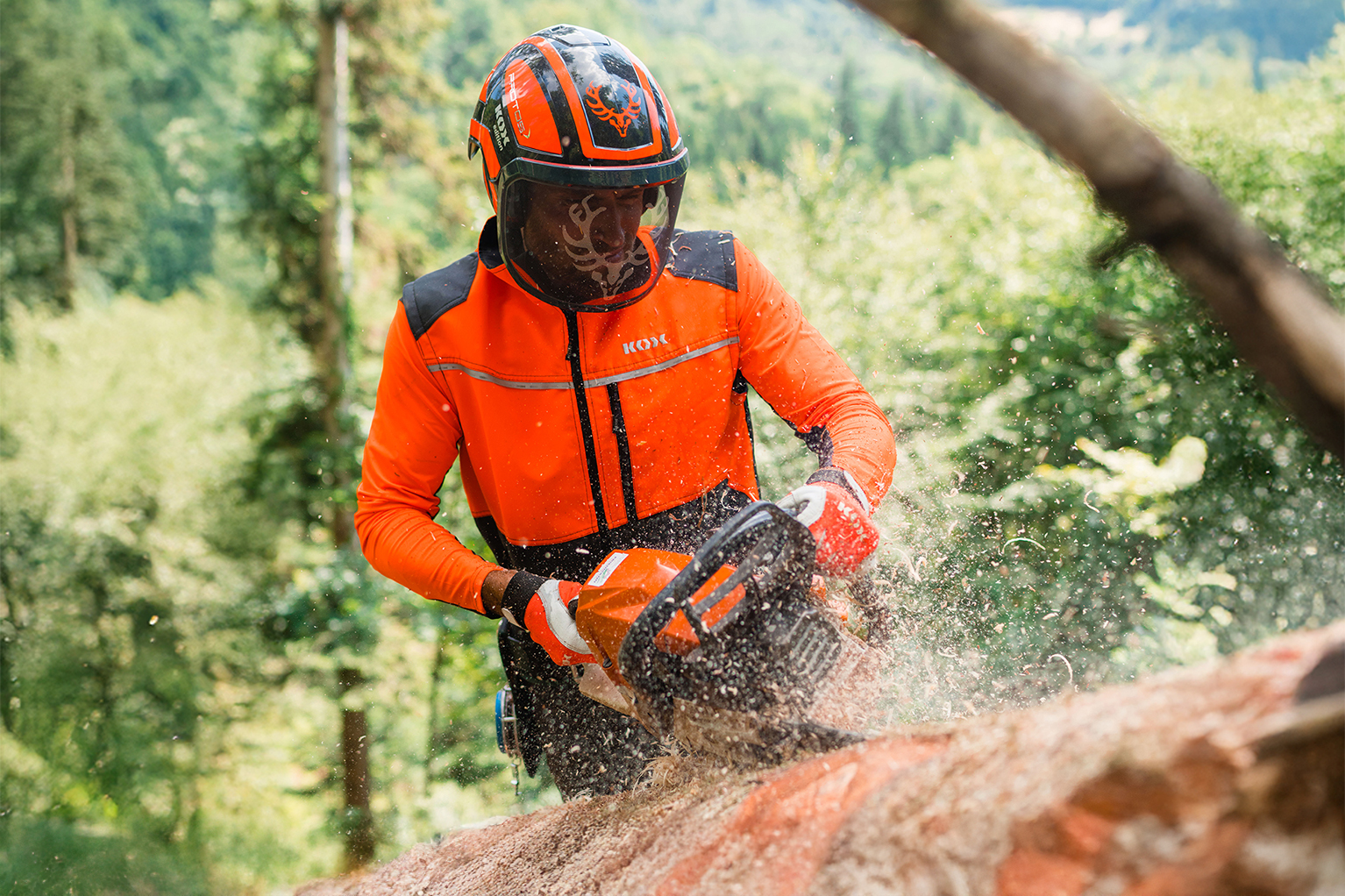 Une personne en tenue de protection foresti�re orange avec casque travaille en for�t avec une tron�onneuse sur un tronc d’arbre, tandis que des copeaux de bois volent dans l’air.