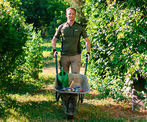 Ein Mann in Arbeitskleidung schiebt auf einem Gartenweg eine Schubkarre mit Gartenger�ten, umgeben von dichtem Gr�n.