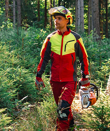 Une personne en tenue de protection foresti�re rouge et jaune avec casque marche dans la for�t en portant une tron�onneuse dans la main gauche.