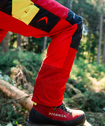 Gros plan sur une personne portant un pantalon anticoupure rouge et noir et une chaussure de travail, avec la jambe fl�chie pos�e sur un tronc en for�t.