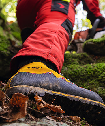 Gros plan sur une chaussure foresti�re robuste � semelle crant�e avec des d�tails jaunes, port�e par une personne en tenue de protection rouge se d�pla�ant sur un terrain irr�gulier en for�t.